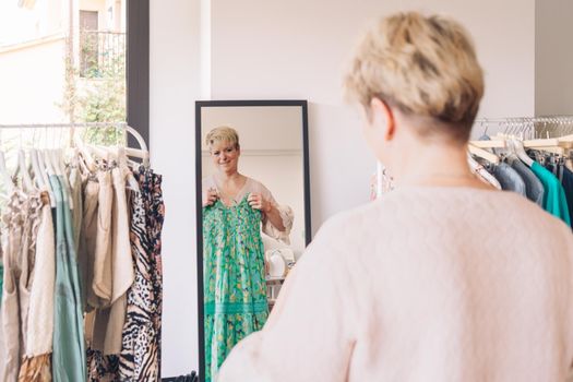 mature woman trying on clothes in front of a mirror. blonde woman shopping in a fashion shop. shopping concept. leisure concept. Natural light, sunbeams, display, clothes rack, clothes, horizontal view, space to copy.