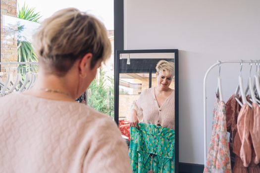 mature woman testing a dress in front of a mirror. blonde female shopping in a fashion shop. shopping concept. leisure concept. Natural light, sunbeams, display, clothes rack, clothes, horizontal view, space to copy.