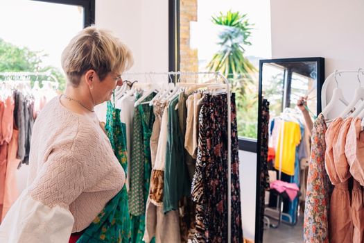 mature woman enjoying a shopping day. blonde woman shopping in a fashion shop. concept of shopping. leisure concept. Natural light, sunbeams, display, clothes rack, clothes, horizontal view, space to copy.