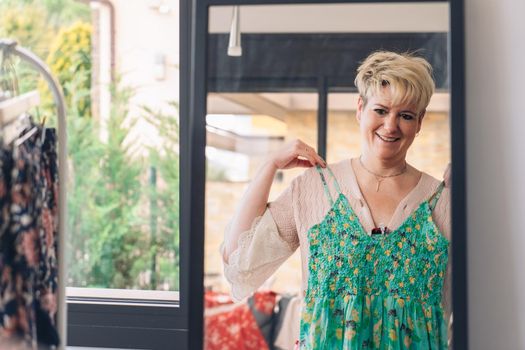 mature woman testing a dress in front of a mirror. blonde female shopping in a fashion shop. shopping concept. leisure concept. Natural light, sunbeams, display, clothes rack, clothes, vertical view, space to copy.