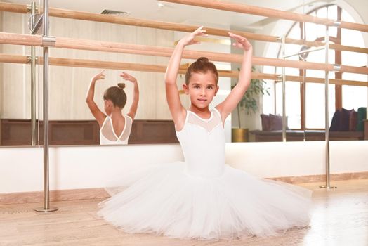 Portrait of a cute little ballerina standing with her hands up in a performance dress. She is dreaming to become a professional ballet dancer. Dance school, ballet studio.