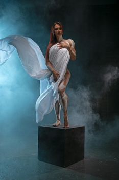 Attractive red-haired girl posing with a piece of white fabric, standing on pedestal like an antique statue in the smoke