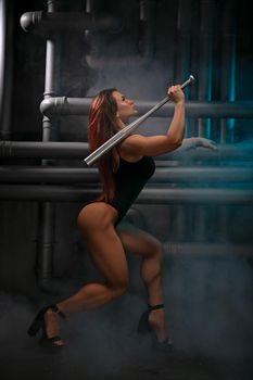 bright girl in black bodysuit with sexy muscular legs in fishnet tights poses with bat in loft studio against the background pipes smoke
