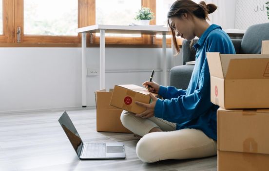 Shipping shopping online, startup small business owner writing address on cardboard box at workplace. Freelance Asian woman small business entrepreneur SME working with box at home.