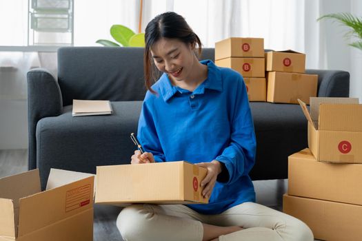 Shipping shopping online, startup small business owner writing address on cardboard box at workplace. Freelance Asian woman small business entrepreneur SME working with box at home.