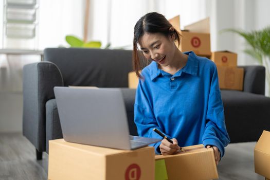Shipping shopping online, startup small business owner writing address on cardboard box at workplace. Freelance Asian woman small business entrepreneur SME working with box at home.