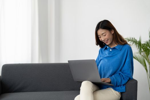 Young asian business woman smile and work from home with laptop computer online to internet on sofa in living room, freelance girl using notebook on couch with comfort, new normal, lifestyle concept.
