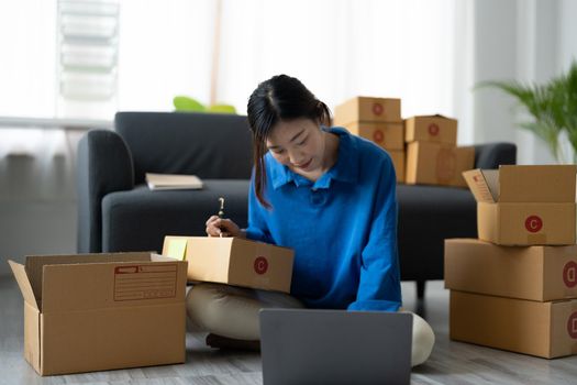 Shipping shopping online, startup small business owner writing address on cardboard box at workplace. Freelance Asian woman small business entrepreneur SME working with box at home.