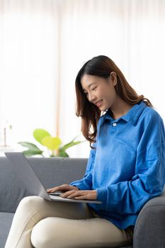 Young asian business woman smile and work from home with laptop computer online to internet on sofa in living room, freelance girl using notebook on couch with comfort, new normal, lifestyle concept.