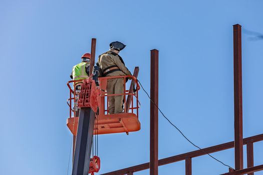 Male builders work at height in a lifting cradle, creating the iron frame of the building.