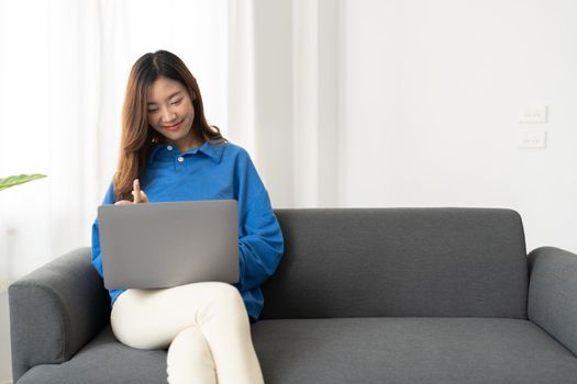 Young asian business woman smile and work from home with laptop computer online to internet on sofa in living room, freelance girl using notebook on couch with comfort, new normal, lifestyle concept with copyspace.