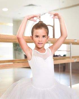 Portrait of a cute little ballerina standing with her hands up in a performance dress. She is dreaming to become a professional ballet dancer. Dance school, ballet studio.