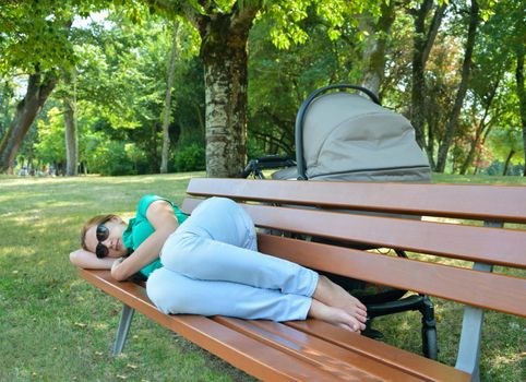 Tired mother asleep on a bench in a park with a baby in a stroller