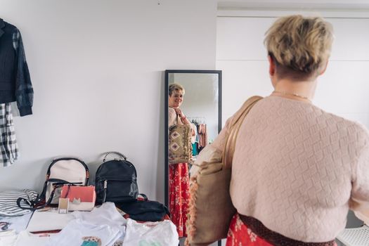 mature woman trying on handbags in front of mirror in fashion shop. blonde woman buying a large handbag. natural light, horizontal view, interior, clothes, hangers. concept of shopping. concept of leisure