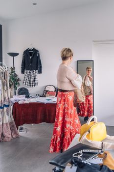 mature woman trying on handbags in front of mirror in fashion shop. blonde woman buying a large handbag. natural light, vertical view, interior, clothes, hangers. concept of shopping. concept of leisure