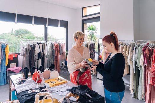 Mature shop assistant charging with credit card. woman paying with contactless in fashion shop. shopping concept. natural light, horizontal view, young woman shopping, mature woman business owner.