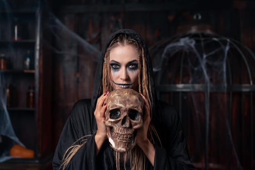 Halloween concept. Witch portrait close up with dreadlocks holding human skull in hand looking camera dressed black hood standing dark room with cage on background Caucasian female necromancer