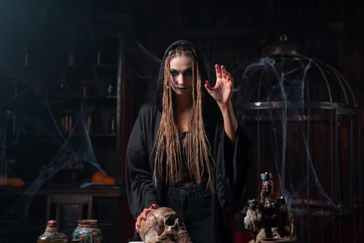 Halloween concept. Witch portrait close up with dreadlocks holding human skull in hand dressed black hood standing dark room side view making curse move hand in camera