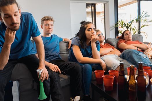 group of disappointed friends watching a football match on television. bored young people at a party at home. concept of leisure. concept of friendship. group of six people, friendly relationship, divided teams, red and blue, competitive.