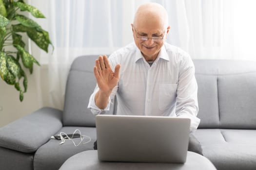 Elderly bald man in white shirt and black pants correcting glasses and working on laptop while sitting in living room