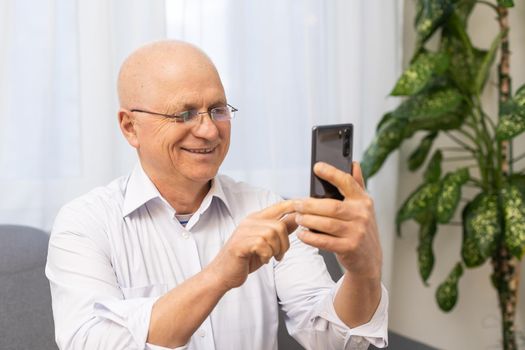 Handsome old man dressed in smart casual style and eyeglasses is using a smartphone and smiling while sitting on couch at home