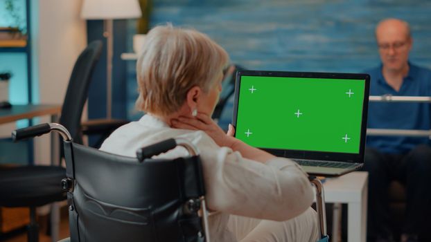 Old person holding laptop with green screen background, sitting in wheelchair. Senior adult with physical disability analyzing blank chroma key with copy space template and isolated mock up.