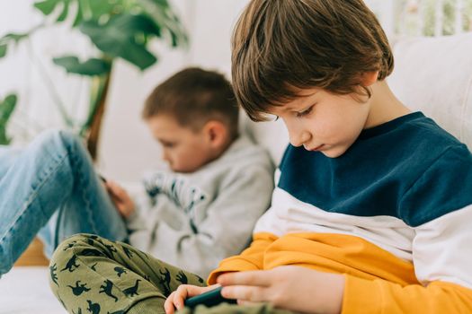 Cheerful two brothers boys playing online game, watching video on cellphone. Smiling kids using funny mobile apps, enjoying free leisure time at home on couch. Children texting together at phone.