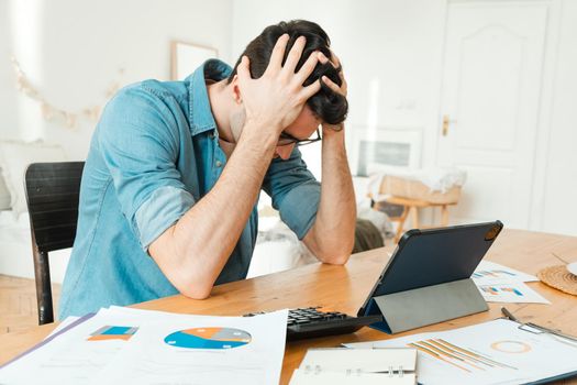 Worried man sitting at the table at home, holding his head, worried about his bills. Loss of money in investments