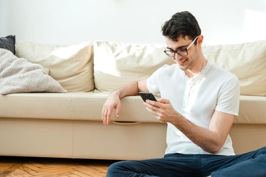 Portrait of an attractive, smiling man in casual clothes sitting on the floor near the sofa in the living room and watching the news using a mobile phone
