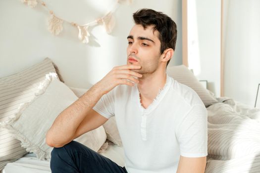 Thoughtful young man sitting on the bed in the bedroom, touching his chin and thinking. Thoughtful mature man resting on bed at home feeling sad thinking about future.