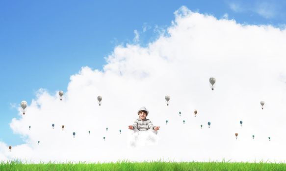 Young little boy keeping eyes closed and looking concentrated while meditating on cloud among flying aerostats with bright and beautiful landscape on background.