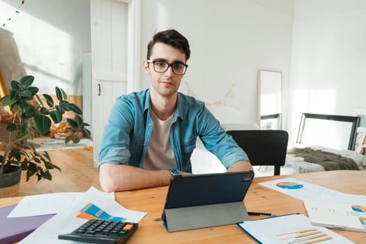 A young man is typing on a laptop, counting on a calculator and working with documents from home. Looking at camera.