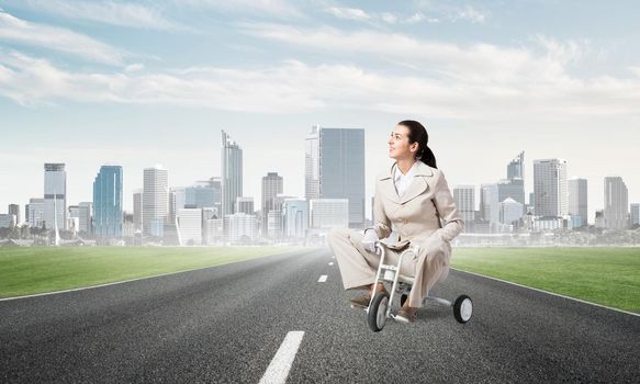 Beautiful young woman riding children's bicycle on asphalt road. Businesslady in white business suit cycling small bike outdoor. Modern city on horizon. Professional career start concept.