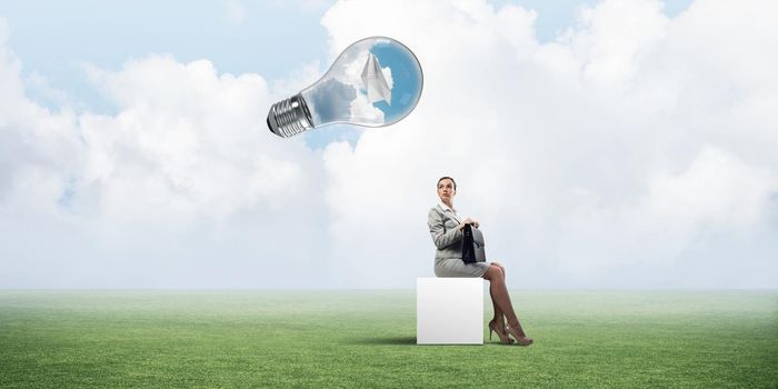 Elegant businesswoman with suitcase in hand sitting on white cube and big glass bulb