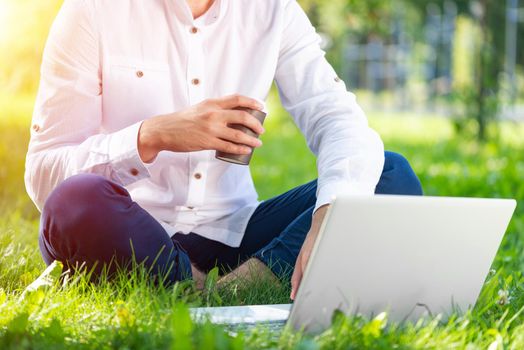 Businessman using laptop computer on green grass in park. Coffee time concept. Close up man hands typing on keyboard. Man working with computer outdoors at sunny summer day. Freelance and blogging.