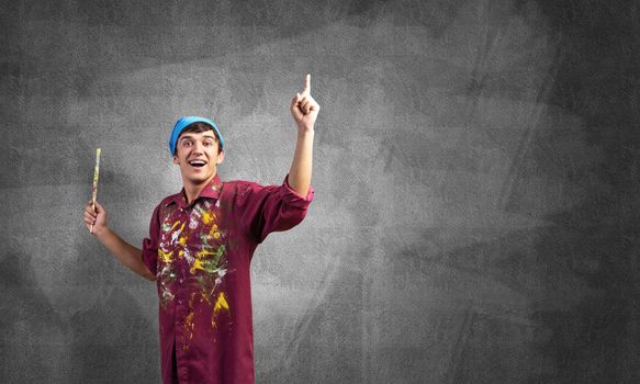 Young emotional artist gesturing with paintbrush. Happy painter in dirty shirt and bandana standing on grey wall background. Creative hobby and artistic occupation concept with copy space