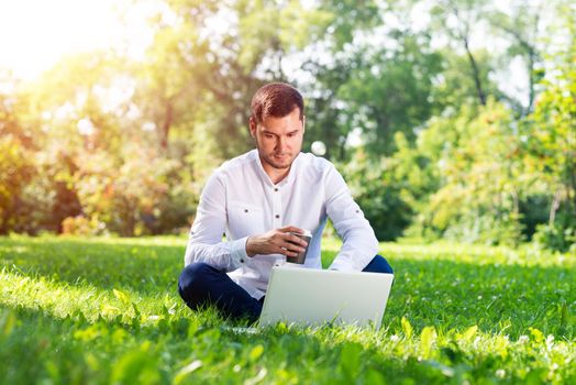 Young businessman with laptop computer on green grass. Coffee break in park. Handsome man in casual wear working with computer outdoors. Enjoying nature at sunny summer day. Remote work concept.