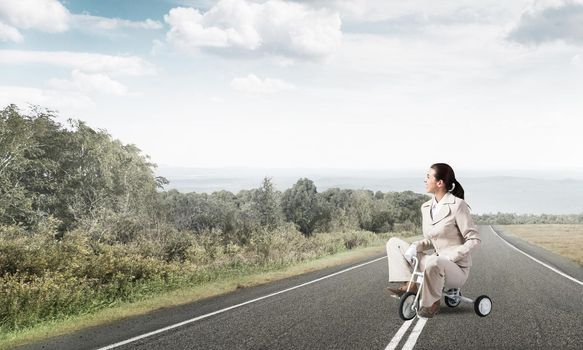 Beautiful caucasian woman riding kid's bicycle on asphalt road. Young employee in white business suit biking outdoor. Professional career start. Beginner level concept with female bicyclist.