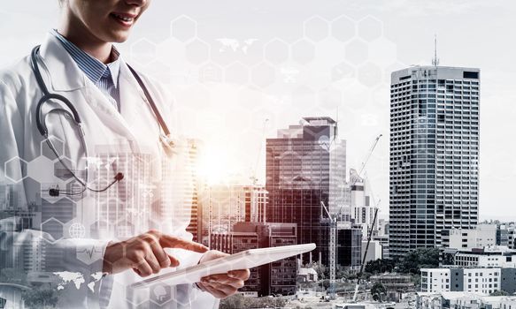 Double exposure of young woman doctor in white medical suit touching tablet with her finger and city view and medical interface on background. Conceptual image of modern medical industry