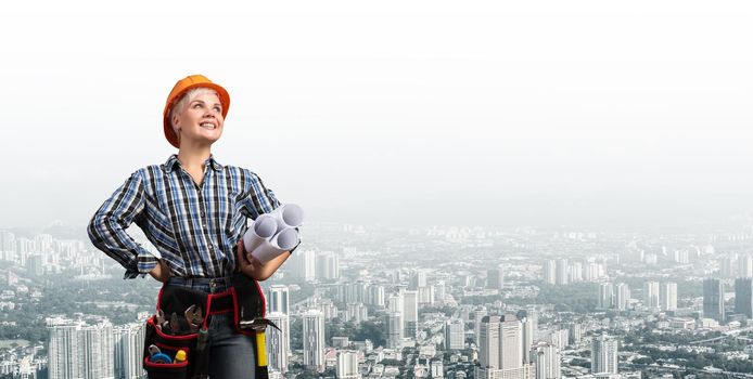 Attractive female engineer in hardhat standing with technical blueprints. Portrait of young architect in checkered blue shirt on background of cityscape. Architecture and construction company.