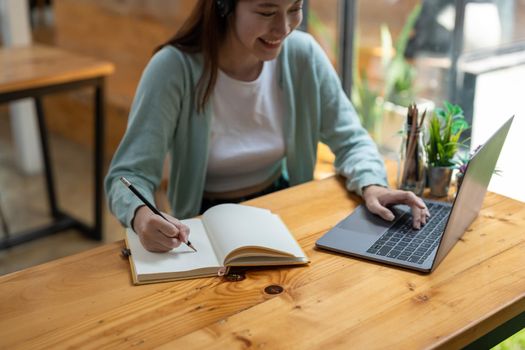 Young Asian girl student wears headphones writing on the notebook to study language online watch and listen to the lecturer, webinar via video call e-learning at home.