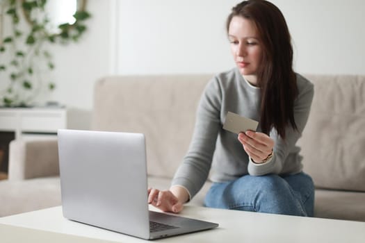 Young woman holding credit card and using laptop at home. Paying online, home shopping, internet banking, electronic store concept.