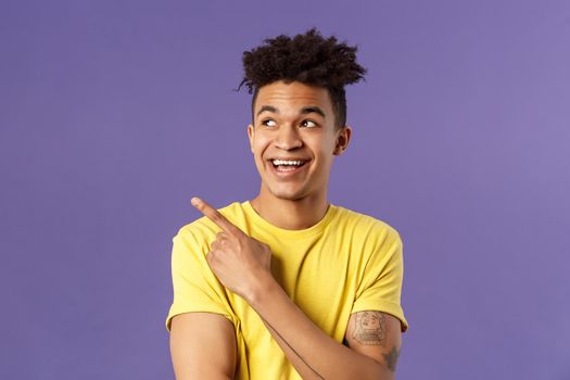 Close-up portrait of funny cute hispanic man with dreads, laughing and smiling at something, pointing looking upper left corner enthusiastic expression, purple background.