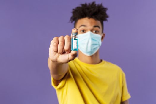 Medicine, coronavirus and people concept. Close-up portrait of young man in facial mask holding ampoule with drug, finally found vaccine from covid19, standing purple background.