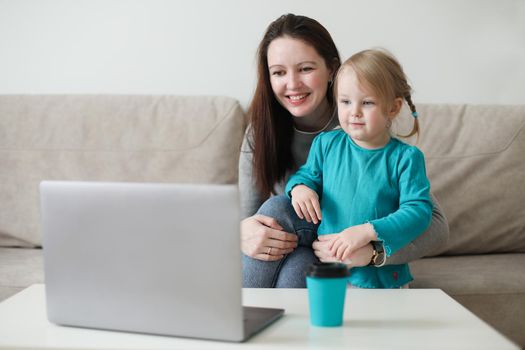 Mother and her daughter using video chat on laptop sitting on cozy sofa at home, watching video, having fun