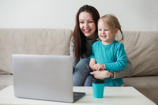 Mother and her daughter using video chat on laptop sitting on cozy sofa at home, watching video, having fun
