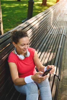 Overhead view of an African smiling sporty woman with smartphone, relaxing on a wooden bench in the city park after hard workout outdoors