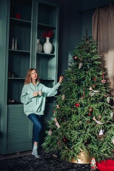 A beautiful woman in homemade clothes near the Christmas tree at home drinks delicious tea or coffee. Festive morning