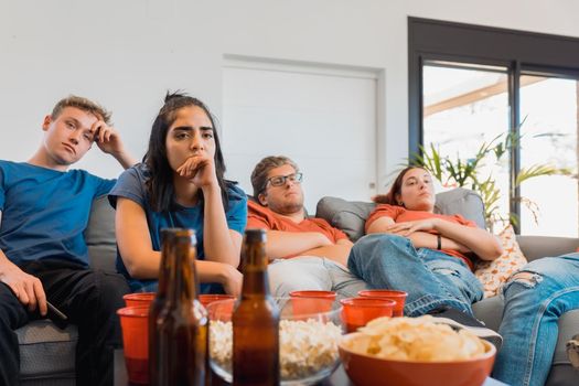 sad friends watching TV because their football team lost the game. upset young people at a house party. concept of leisure. concept of friendship. group of six people, friendly relationship, divided teams, red and blue, competitive.