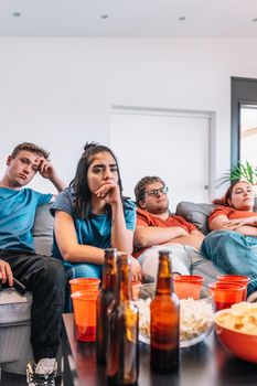 sad friends watching TV because their football team lost the game. upset young people at a house party. concept of leisure. concept of friendship. group of six people, friendly relationship, divided teams, red and blue, competitive.